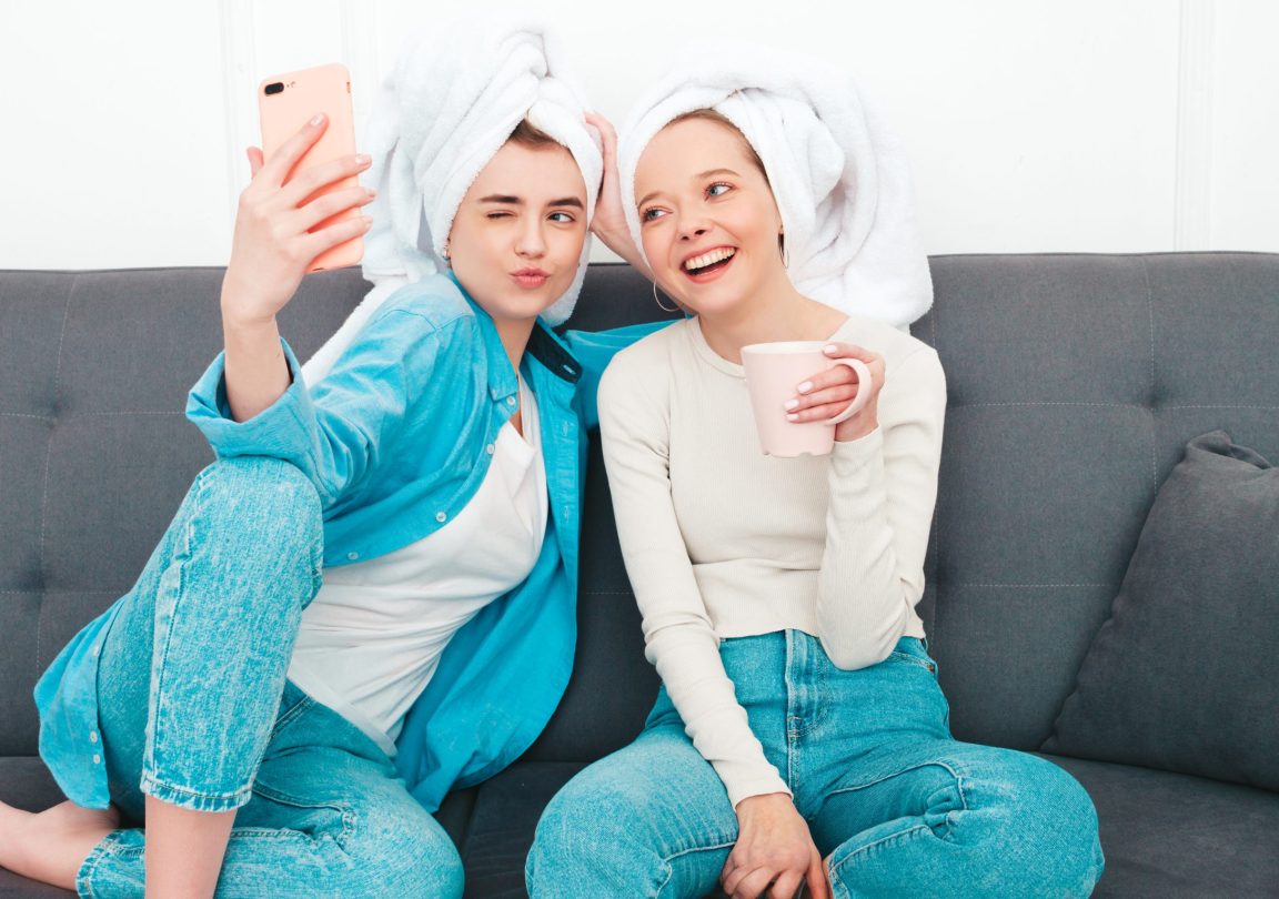 Two young beautiful smiling women sitting at the sofa. Sexy carefree models posing indoors in posh apartment or hotel room. They doing beauty treatments at home in towels on heads.Taking selfie photos
